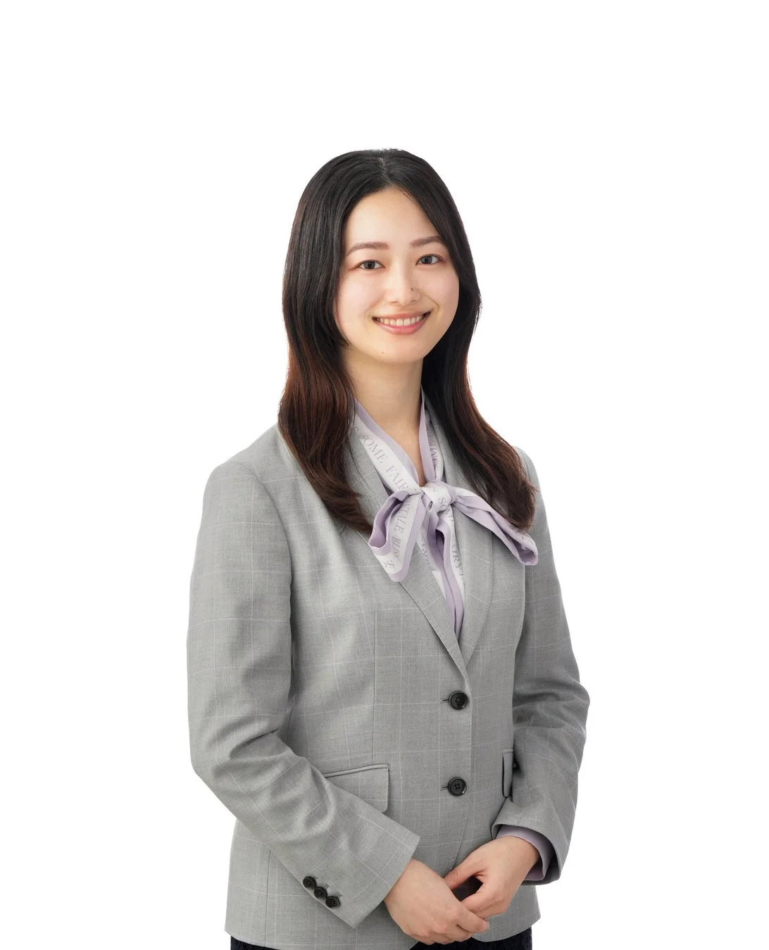 白い背景の前で、グレーのチェック柄スーツと薄紫色のスカーフを身につけた若い女性が笑顔で立っているポートレートです。プロフェッショナルで好印象を与えます。