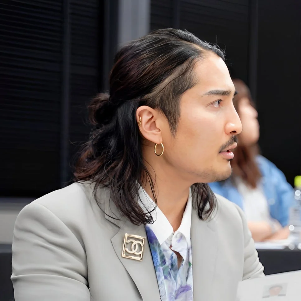長髪を束ねた男性が、グレーのスーツジャケットに花柄のシャツを着用し、シャネルのブローチをつけている。横顔が写っており、会議のような屋内の場面で何かを見ている様子。