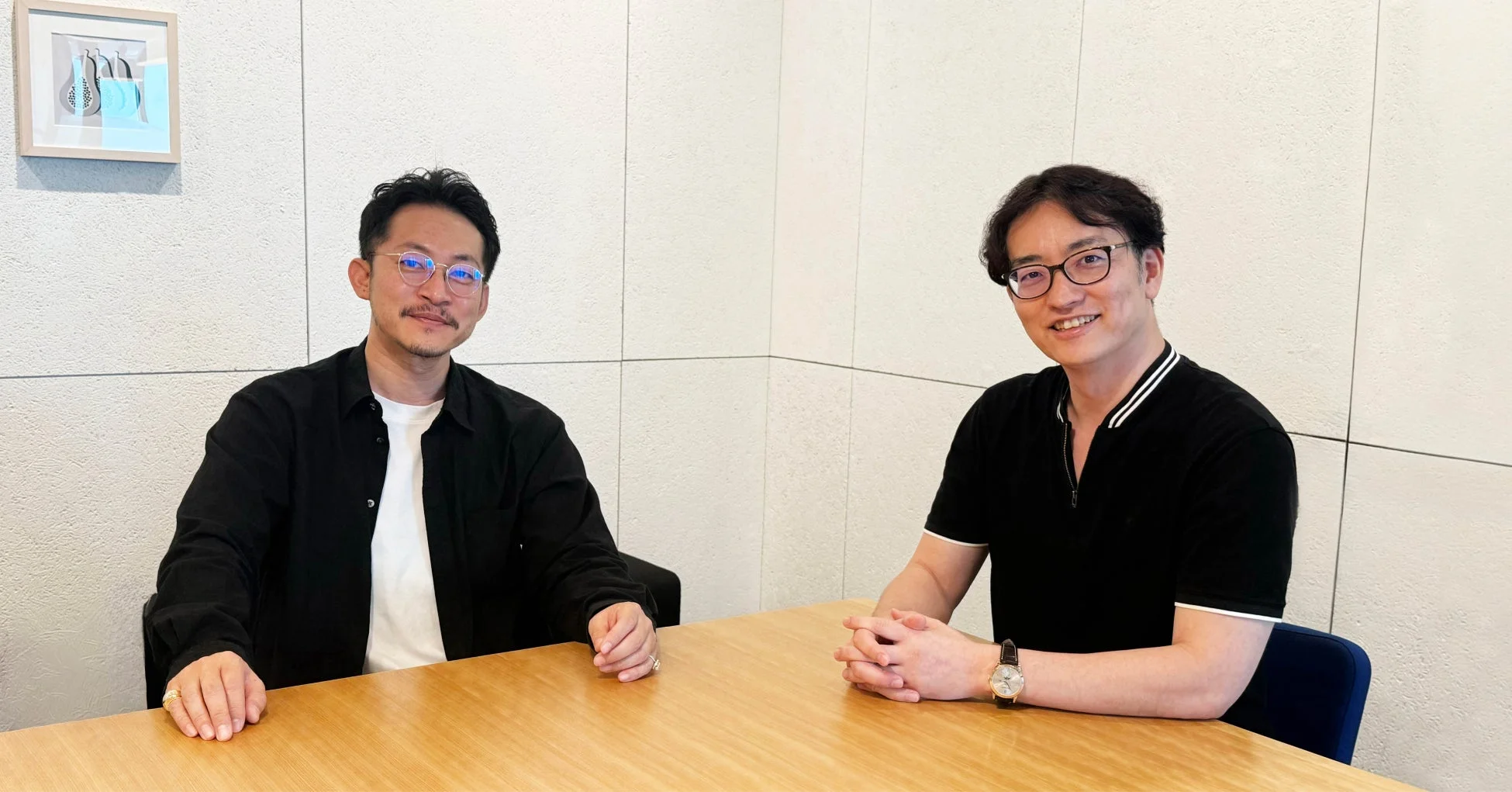 木製のテーブルに座ってカメラに笑顔を向ける2人の男性。カジュアルな服装で、対談や会議の様子を思わせる一枚です。